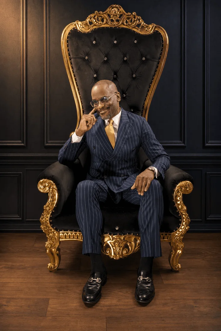 Dr. Glenn Toby in a navy pinstripe suit, seated on a black and gold throne against dark paneled walls